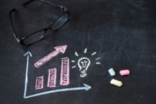 Eine Brille liegt auf einer bemalte Tafel, auf der ein Säulendiagramm und eine Glühbirne aufgezeichnet sind. Es lieben drei farbige Kreiden auf der Tafel.