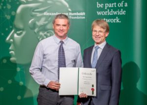 Two people are standing in front of an advertising banner of the Humboldt Foundation with a certificate in their hands and are looking into the camera.