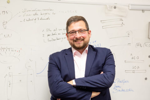 A smiling man with glasses stands with arms crossed in front of an inscribed blackboard.