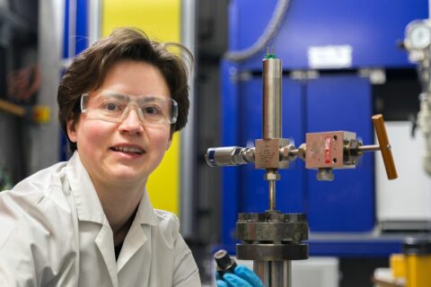 A person with transparent goggles and a white lab jacket looks into the camera. In the background you can blur a large plant in blue and yellow color.