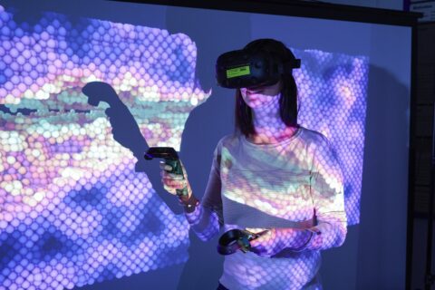 A person has VR glasses on and stands in front of a colorful wall.