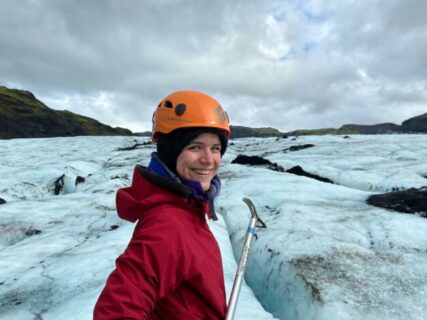 Nora is standing on a glacier, wearing a red jacket and an orange helmet on her head.