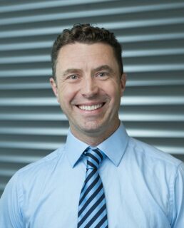 A person with short, brown hair smiles kindly at the camera. The person is wearing a light blue shirt and a blue and black striped tie.