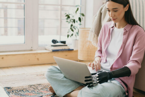 Close-up picture of beautiful charming female in pale pink silk shirt sitting on floor on colorful carpet holding laptop on knees with prosthetic bionic hand made of black metal mechanical device