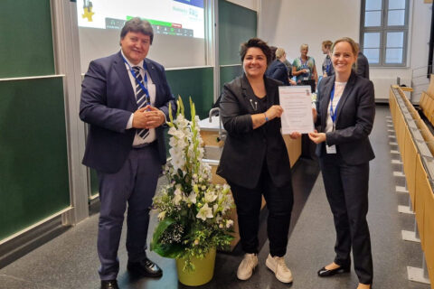 3 people stand next to each other and look into the camera. They are standing in front of the rows of seats in a lecture hall, next to them is a flower pot with large, white flowers.