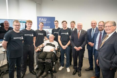 Several people in blue T-shirts or suits stand in a semi-circle, including one person in a wheelchair.