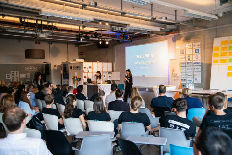 In a room, many people sit on chairs and look at a person who is giving a lecture.