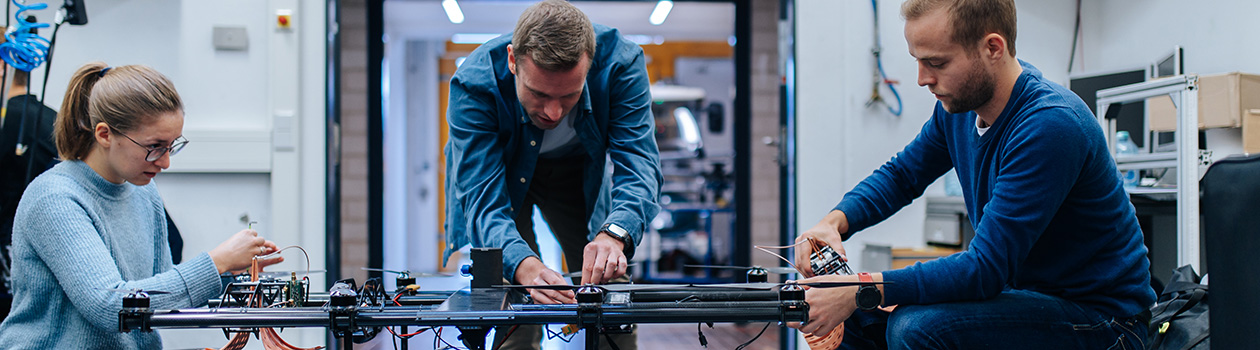 3 people sit around a large suitcase with metal fasteners. The people are each working with both hands on a flying drone standing on the suitcase.