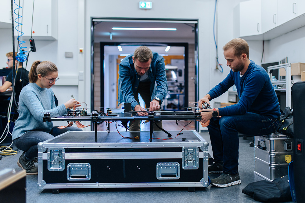 3 people sit around a large suitcase with metal fasteners. The people are each working with both hands on a flying drone standing on the suitcase.