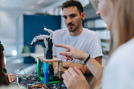 Two people are looking at a mechanical hand. In the foreground, a blurred woman can be seen pointing at the hand, in the background a man watching her. The mechanical hand is connected with colorful cables.