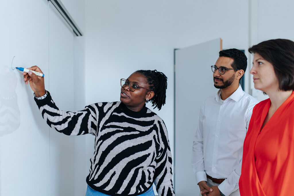 One person stands in front of a whiteboard, writing on it with a blue pen. Two other people are looking at the whiteboard.