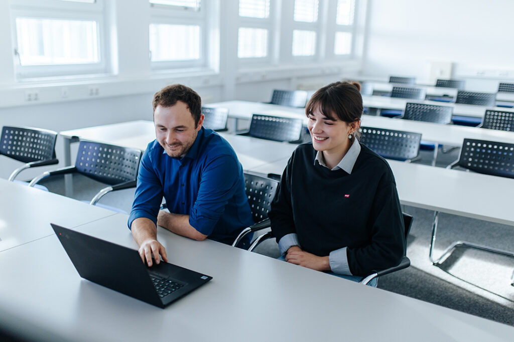Two people are sitting in a large room with rows of tables and chairs. They are looking into a laptop.
