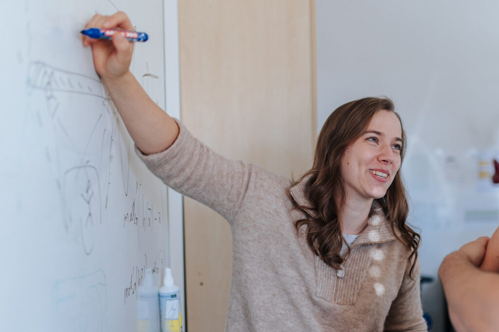 A woman is standing in front of a whiteboar while explaining something to a person outside the picture.