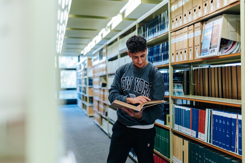 A male student is standing between the shelves of a library. He is reading a book.