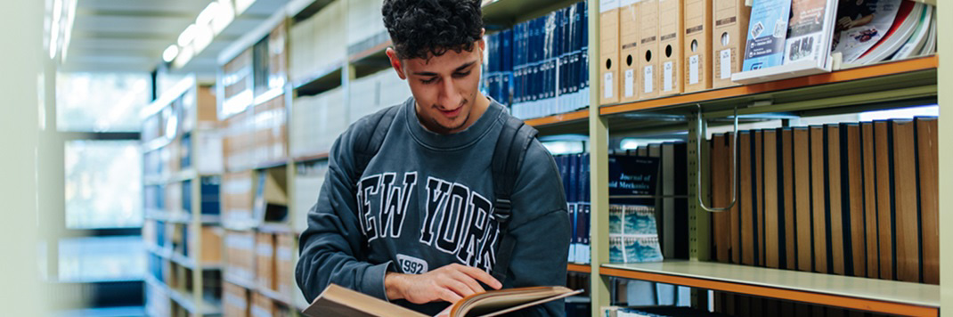 A male student is standing between the shelves of a library. He is reading a book.