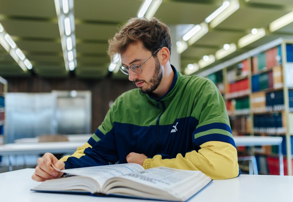 A young man wearing a green jacket is reading a book in a library.