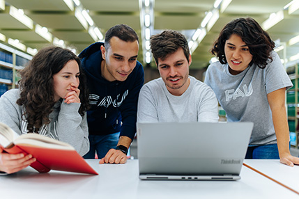 Four students are sitting in a library looking at the screen of a laptop.