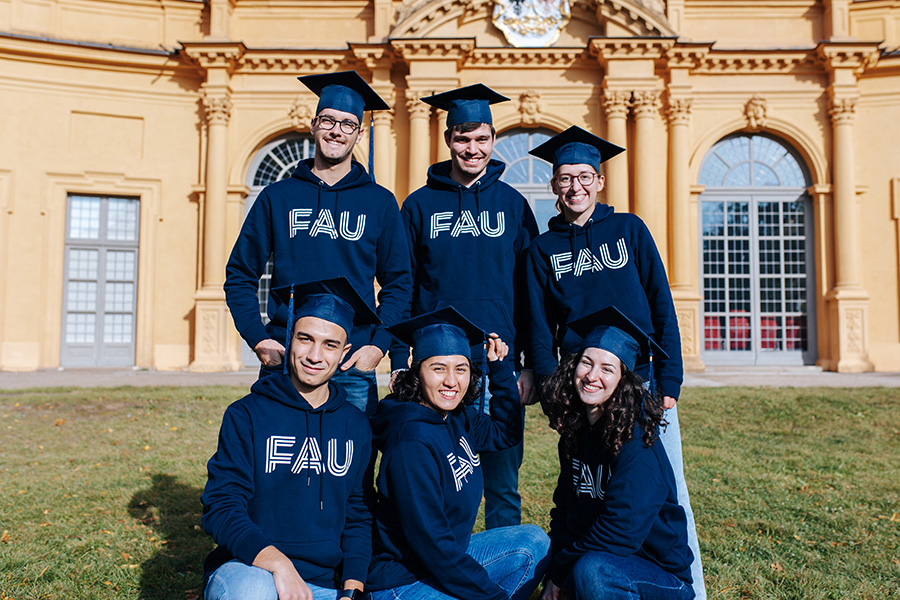 A group of six people are standing in front of an orange building with large windows. They are all wearing blue graduate hats and a blue hoodie with FAU printed on it.