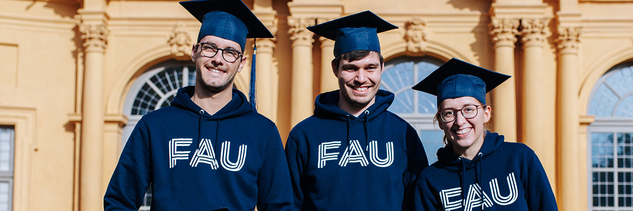 Three people are standing in front of an orange building with large windows. They are all wearing blue graduate hats and a blue hoodie with FAU printed on it.