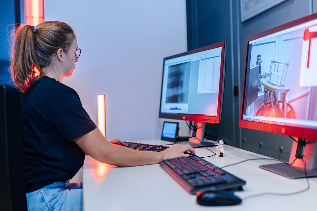 A woman with glasses and a braid is sitting at a desk with two computer screens and two computer keyboards. The woman is looking at one of the screens and typing on the keyboard.