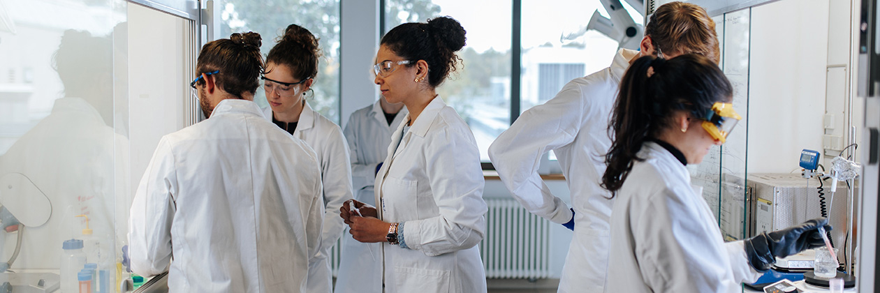 Several people wearing protective goggles and white coats are standing in a laboratory, talking and working together.