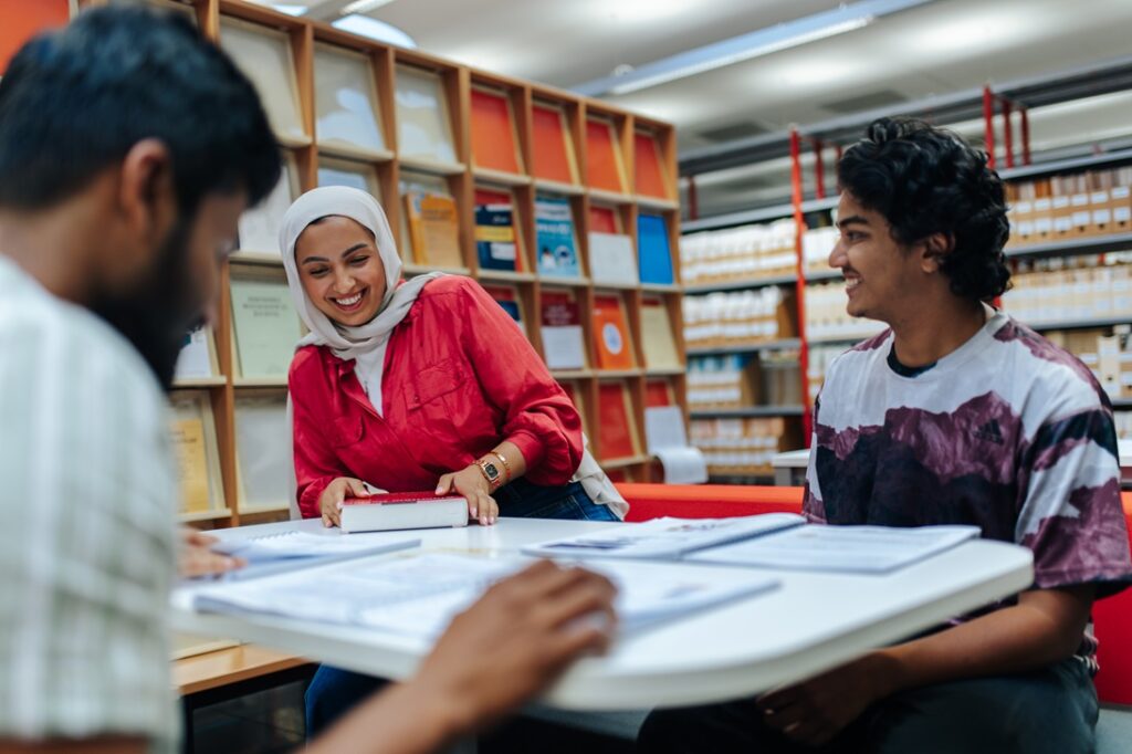 Three students are sitting in a library and are working and laughing together.