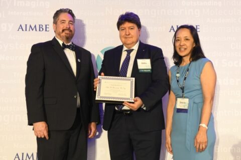 2 men in dark suits and a woman in a light blue dress stand next to each other in front of a white wall. One person is holding a certificate in their hands, all are smiling friendly into the camera.