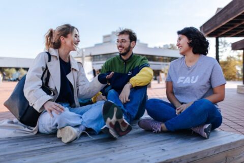 A group of three young people are sitting on a wooden bench in a square in front of a building.