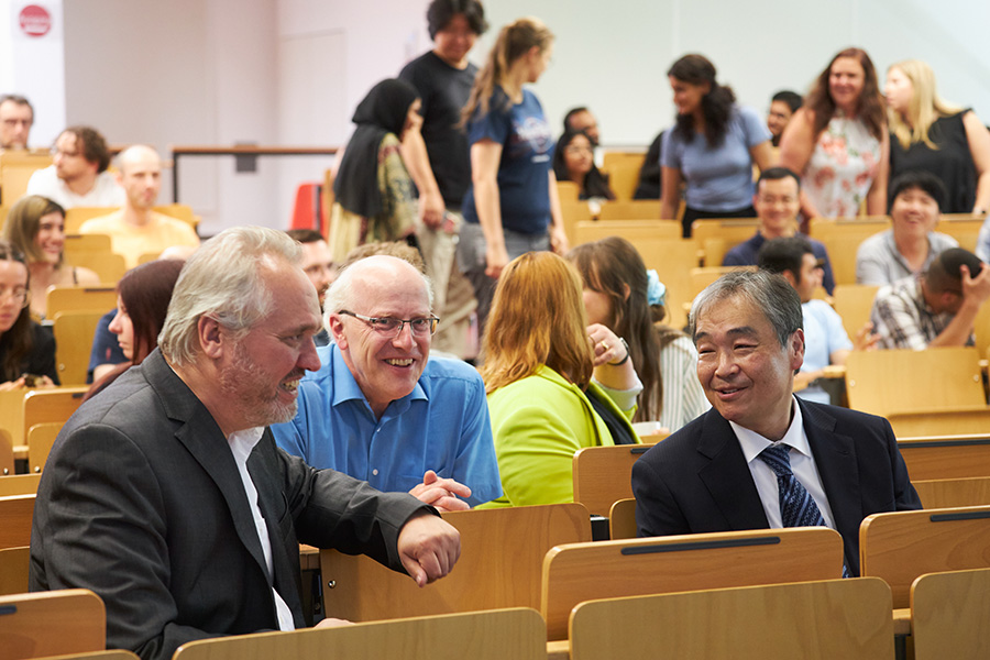 In a room with many rows of seats, several people are sitting and standing. The focus is on three people in the front row, these people are talking to each other with friendly smiles.
