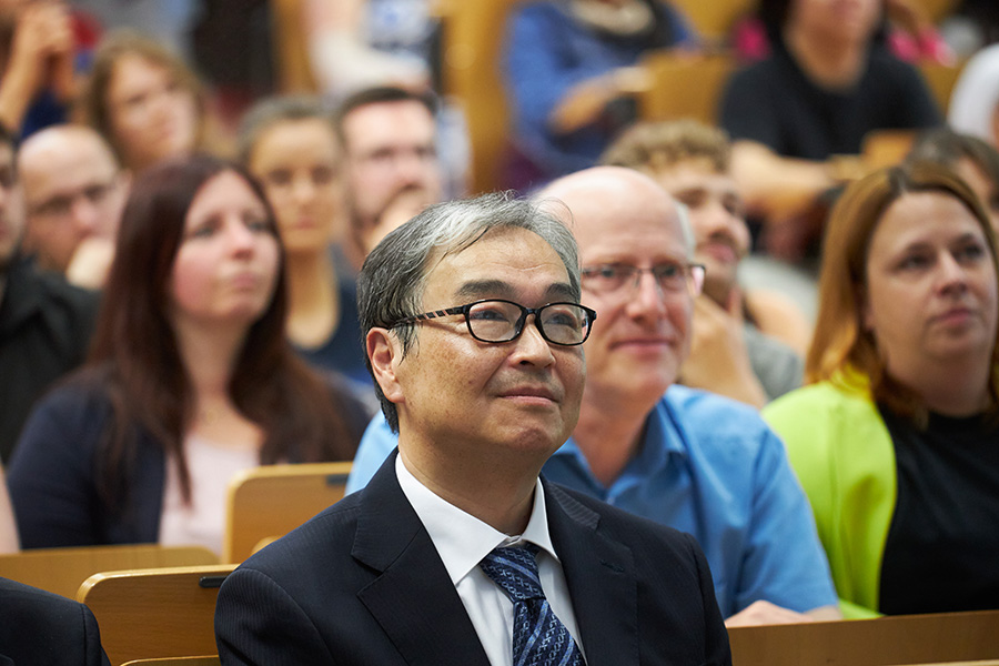 A person can be seen in the foreground wearing a dark suit with a white shirt and dark tie. The person wears glasses, looks forward in a friendly manner and sits in a row of chairs in front of other people.