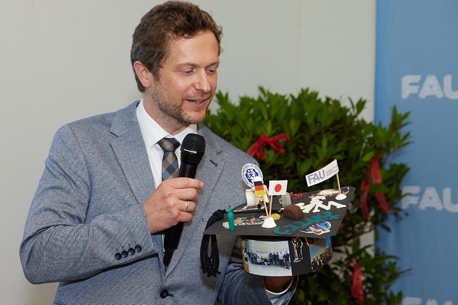 A person in a light-colored suit holds a doctor's hat in one hand and a microphone in the other.