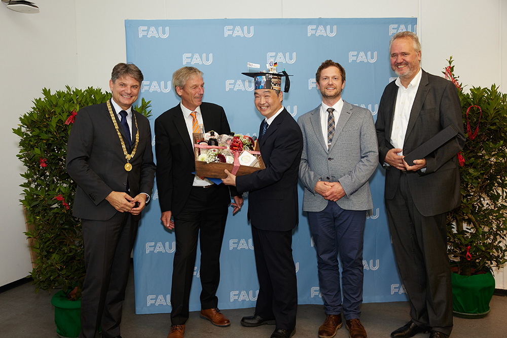 5 people stand in front of a light blue wall on which FAU is written in white letters. They are all smiling and looking at the camera. The person in the middle is wearing a doctor's hat and holding a gift basket.
