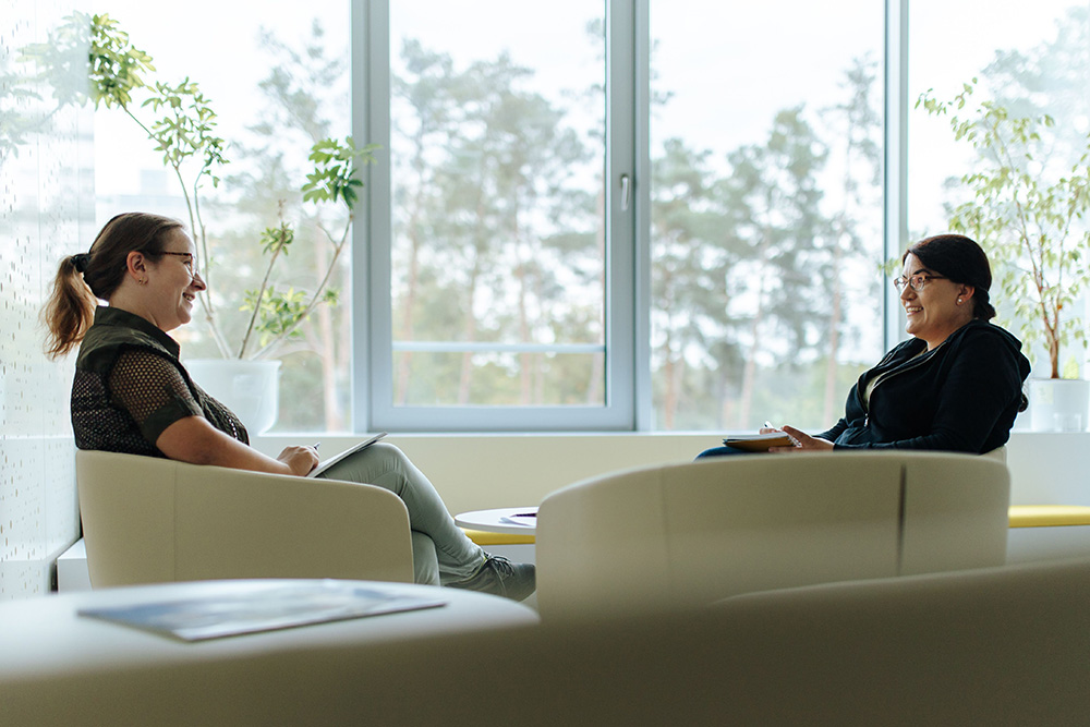 Two people sit opposite each other in armchairs in front of a large window. They are both holding a pen and paper and looking at each other in a friendly manner. They both have dark hair, are wearing a dark top and glasses.