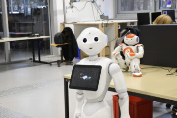 A white robot with a black screen on its chest stands in front of a table and looks into the camera with a friendly face. A small, white and orange robot is sitting on the table.