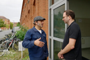 Two people are standing at the front door of a red building. They are talking to each other. Bicycles can be seen in the background.