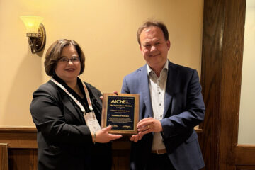 Two people are standing next to each other, holding a framed picture in front of them. They are looking at the camera with a friendly expression.