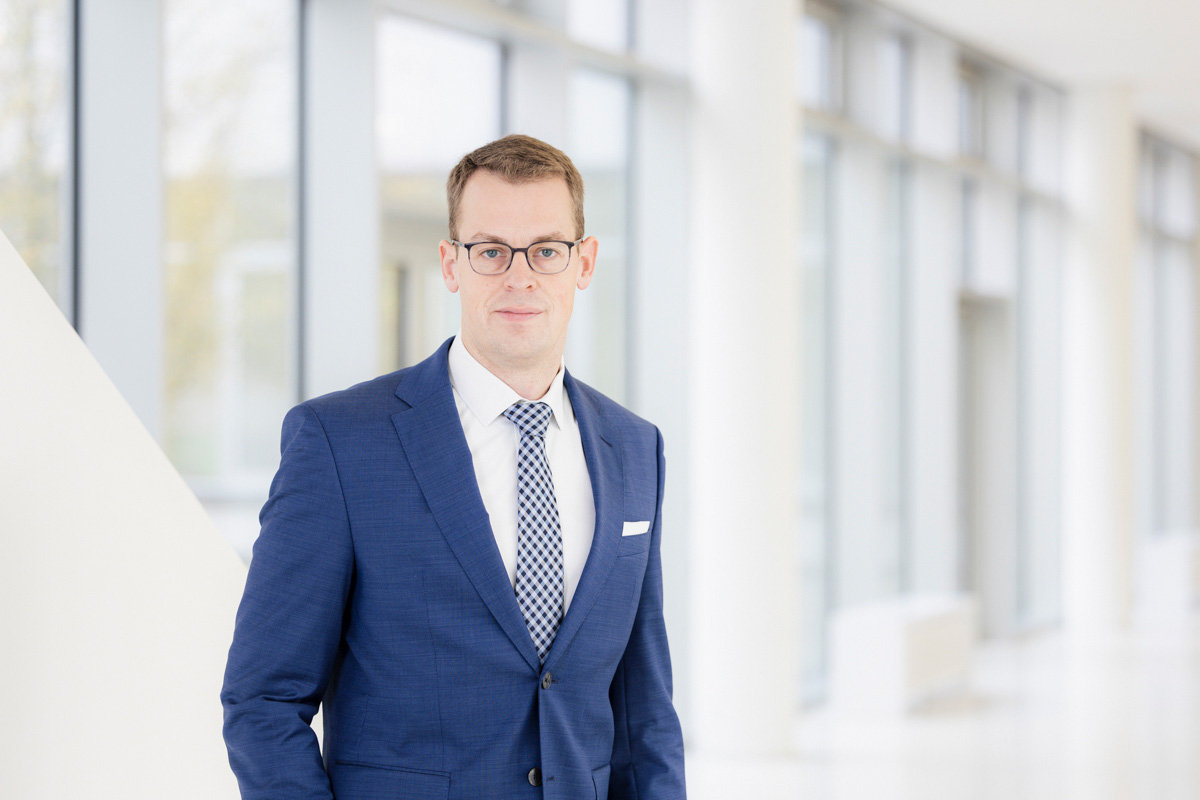 A person with short brown hair and glasses looks into the camera. The person is wearing a white shirt with a blue and white checkered tie and a blue jacket over it.