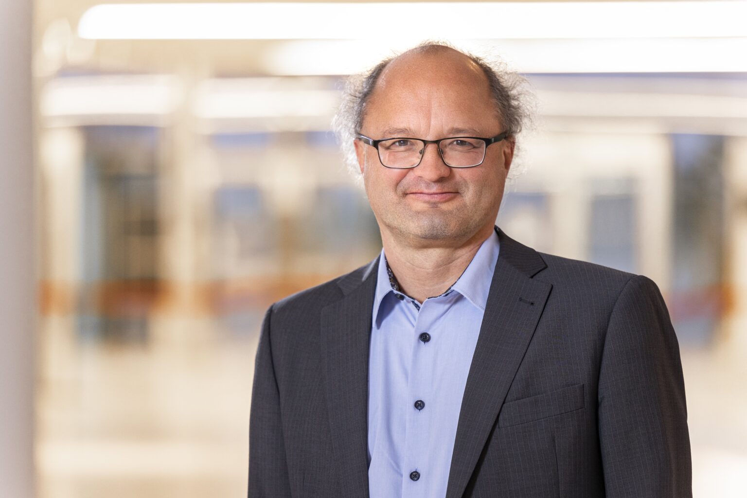 A person wearing glasses, a blue shirt, and a dark jacket is smiling at the camera.