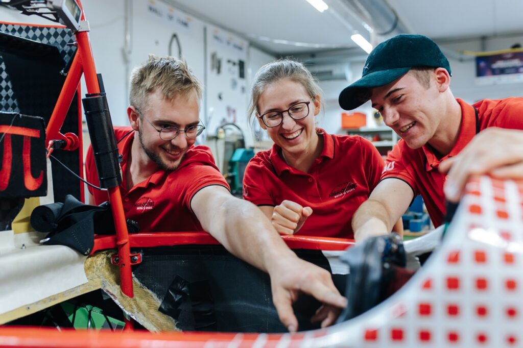 A group of two young men and a young woman are working on a project in a workshop. They are wearing red T-shirts. The man on the left is pointing at the car they are currently working on.