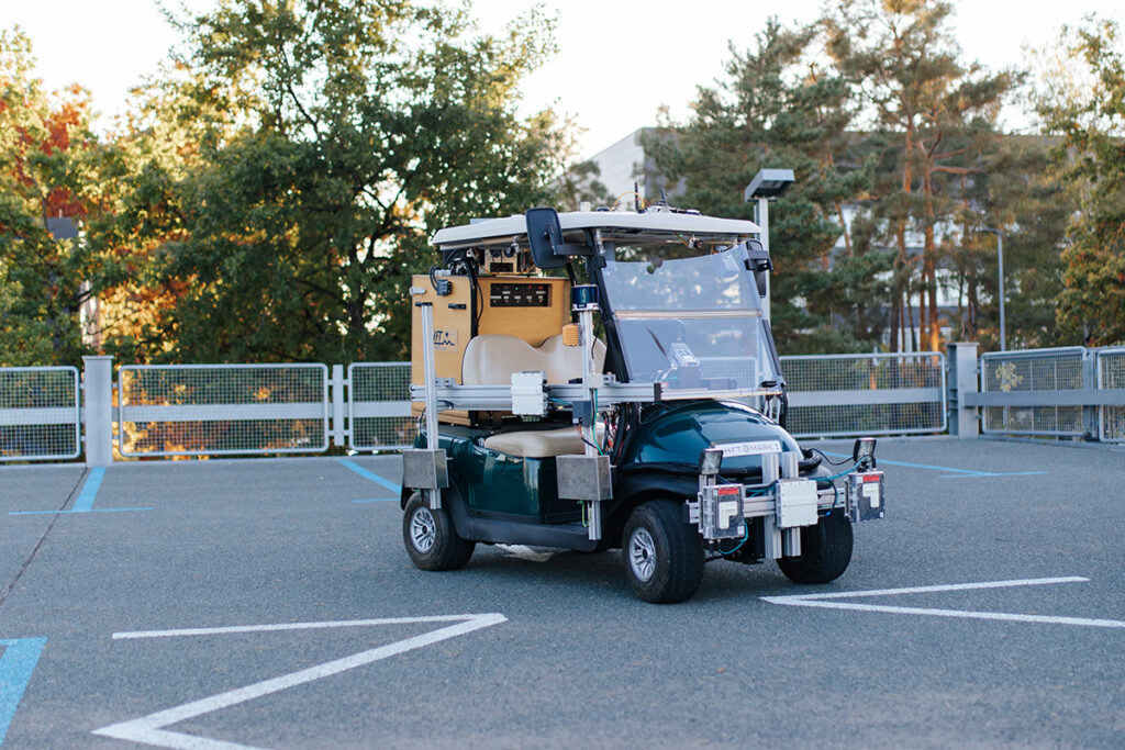 A golf cart is parked in a parking lot with equipment attached to it for research into autonomous driving. Trees and a building can be seen in the background.