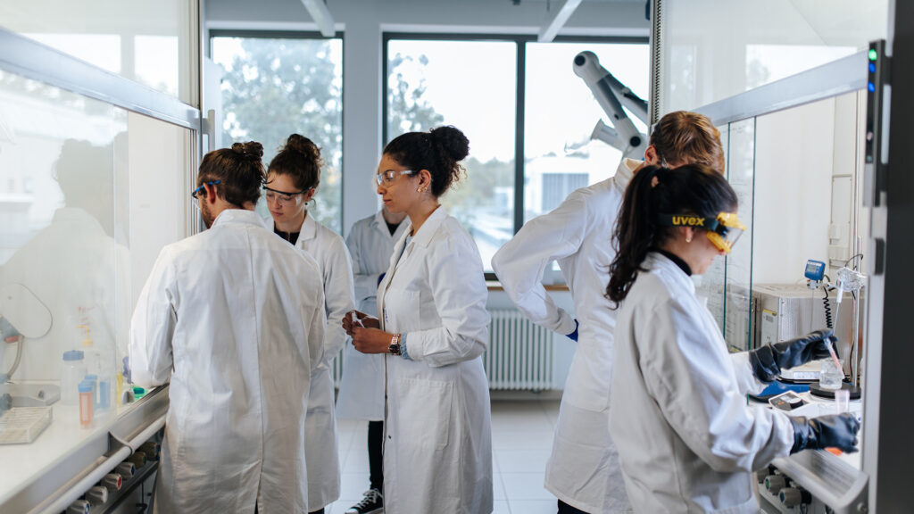 Several people wearing protective goggles and white coats are standing in a laboratory, talking and working together.